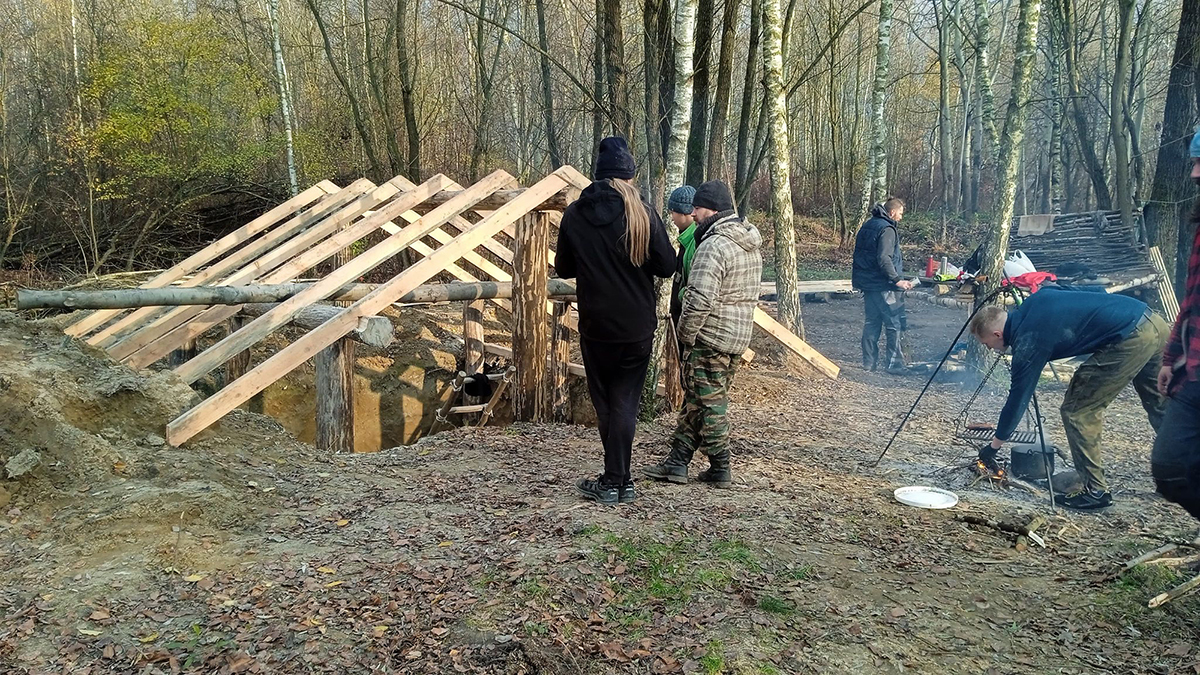 etap budowy drewnianej chaty. z ziemia wyłania się konstrukcja dachu. wokół krzątają się ludzie