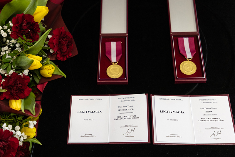 Dwa złote medale z amarantowymi wstążkami, pod nimi dokumenty potwierdzające ich nadanie