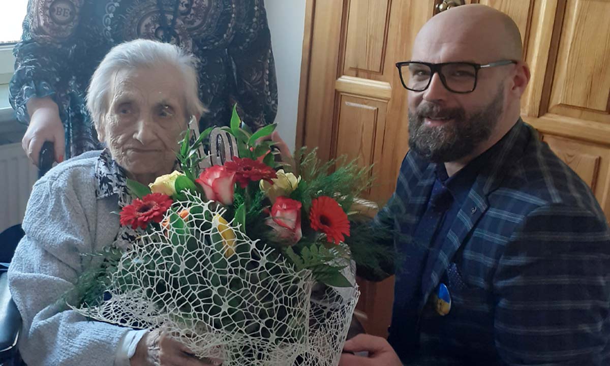 Starsza kobieta z bukietem kwiatów na wózku inwalidzkim, obok mężczyzna w garniturze, okularach i z brodą. 