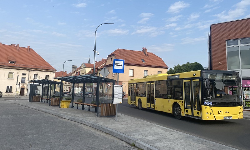 Dworzec autobusowy w otoczeniu kamienic, na pierwszym planie ruszający żółty autobus