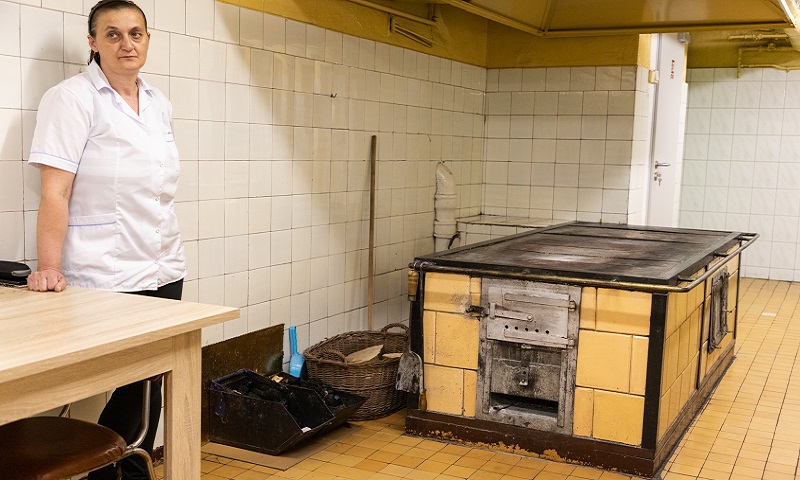 Pomieszczenie kuchenne, po lewej stronie kobieta białym fartuchu, po prawej stary, kuchenny piec na węgiel