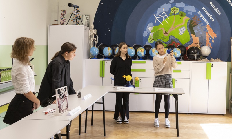 Sala szkolna, na pierwszym planie uczniowie przy stołach wykonują eksperyment, za nimi interaktywna plansza z budową stratosfer ziemi
