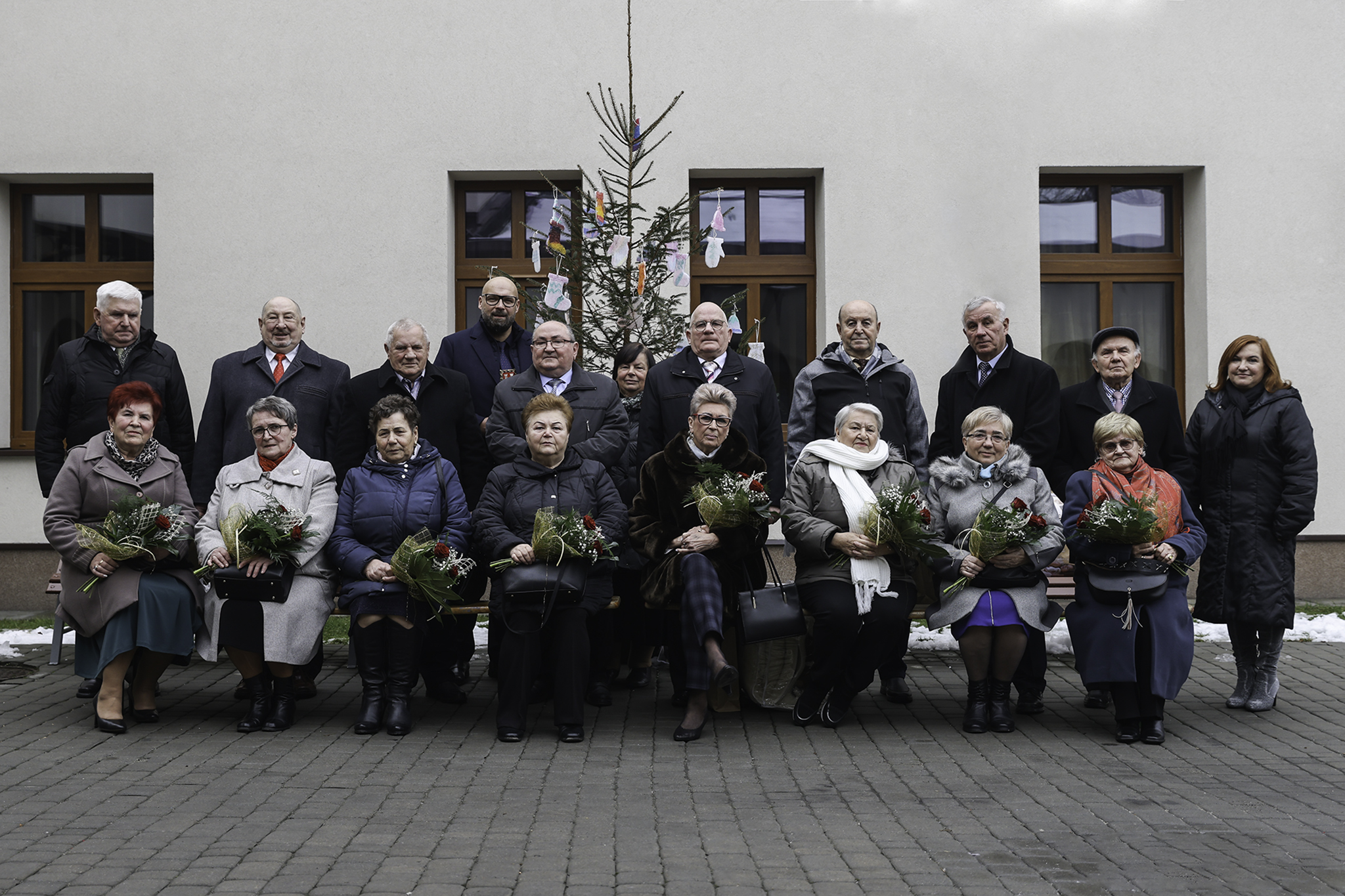 Grupa osób, kobiet i mężczyzn, przed białym budynkiem. Kobiety siedzą na krzesłach, trzymają na w rękach kwiaty. Za nimi żywa choinka