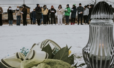 Na pierwszy planie znicz, w tle zaśnieżony plac i grupa młodzieży w okręgu, w zimowych ubraniach. Za nimi linie elektryczne kolei