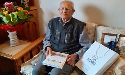 Starszy mężczyna w koszuli i swetrze siedzi na kanapie. Na kolanach trzyma książkę Rocznik Pyskowicki, po lewej stronie flakon z bukietem kwiatów, w tle meblościanka. 