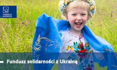 Dziewczynka w wianku, z blond włosami, w stroju ludowym, pośród zbóż. Podpis Fundusz Solidarności z Ukrainą