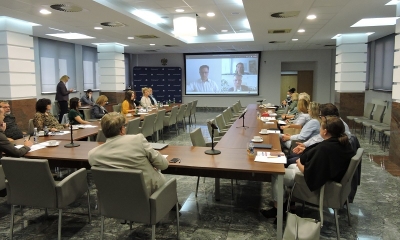 Ludzie oficjalnie ubrani siedzą wokół stołu konferencyjnego z głowami zwróconymi w stronę ekranu, z którego przemawia prelegent