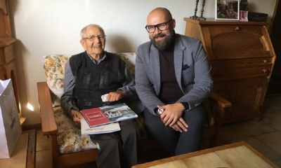 Dwóch mężczyzn na kanapie, z lewej w bardzo podeszłym wieku, obok dużo młodszy, z brodą i w okularach. Uśmiechają się do obiektywu