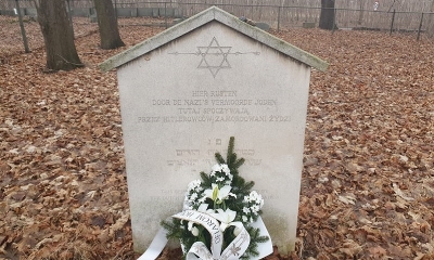 Macem żydowska wsród liści  i innych nagrobków, przed nią wiazanka kwiatów, białych lilii. 