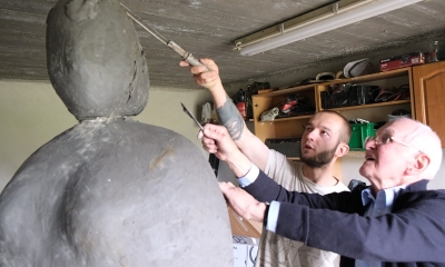 Dwóch mężczyzn, młody i senior, pracują nad rzeźbiarskim, kulistym obiektem. Młodszy trzyma w rękach dłuto 