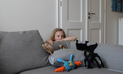 Dziewczynka o kręconych blond włosach schowana za szarą kanapą, na której leżą pluszaki - potwory