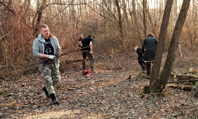 Zdjęcie lasu, trzech mężczyzn porządkuje teren, ten na pierwszym planie ubrany w dżinsową kurtkę niesie pęk gałęzi