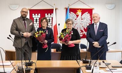 Zdjęcie zbiorowe, na tle herbu Pyskowic, godła i flagi Polski. Z prawej i lewej strony mężczyźni w garniturach, w środku dwie kobiety uhomorowane medalami, z bukietami w rękach
