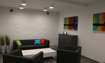 Pomieszczenie z wybiałkowanymi ścianami. Pod nimi kanapy, na jednej z nich bardzo kolorowy obraz