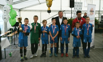 Siódemka młodych chłopców w niebieskich strojach piłkarskich w namiocie, na szyi medale, chłopiec po lewej trzyma puchar. Za nimi dwóch mężczyzn - jeden w garniturze, drugi w dresie, trzymają ręce na ramionach chłopców. 